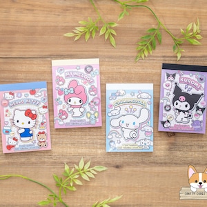 May include: Four colorful memo pads featuring popular cartoon characters, including Hello Kitty, My Melody, Cinnamoroll, and Kuromi. Each pad has a different color scheme and character design, with the characters in various poses. The pads are arranged on a wooden surface with green foliage.