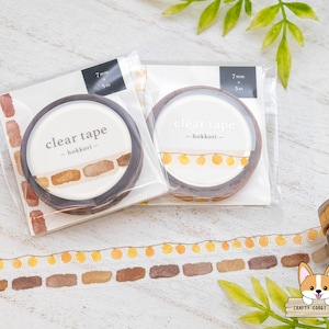 May include: Two rolls of clear tape with a brown background and the text "clear tape -hokkori-." The tape is decorated with a pattern of brown rectangles and yellow circles. The tape is on a white wooden surface.