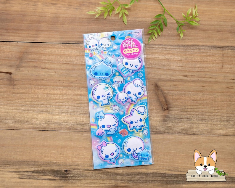 May include: A package of stickers featuring cute, cartoonish characters in various poses. The stickers have a blue and pink color scheme with a rainbow and star design. The package has Japanese text and a pink circular sticker.