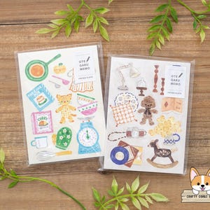 May include: Two clear plastic packages of paper stickers with various illustrations. One features cooking-themed images like a pan, scale, and cat. The other includes a lamp, teddy bear, and rocking horse. Both have the text "OTE GARU MEMO PAPER PLATES".