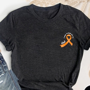 Peut inclure: T-shirt gris foncé avec un ruban orange et le texte "I wear orange for my dad". Le t-shirt est fait d'une matière douce et a un design classique à col rond. Le t-shirt est noué en bas.