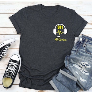May include: A dark gray t-shirt with a white graphic of a headset with a heartbeat and the text "911 Olivia" in yellow.