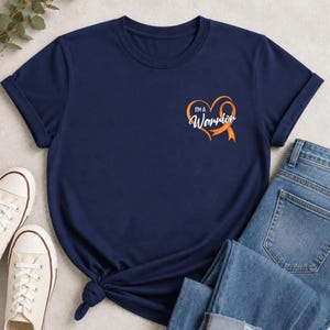 Può includere: Una t-shirt a maniche corte blu navy con una grafica sul lato sinistro del petto. La grafica consiste in un nastro arancione di sensibilizzazione a forma di cuore con il testo bianco "I'M A Warrior" in un carattere corsivo.