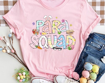 Può includere: Maglietta rosa con una grafica colorata che dice "Para Squad" con elementi a tema pasquale come orecchie di coniglio, uova e un cestino.