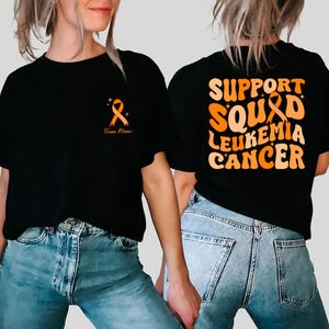 Peut inclure: T-shirts noirs avec des graphiques de rubans orange. Un t-shirt porte un ruban et le texte "Team Name". L'autre t-shirt porte les mots "SUPPORT SQUAD LEUKEMIA CANCER" en orange. Les t-shirts sont portés avec des jeans bleus.