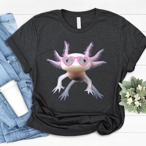 May include: A dark grey t-shirt featuring a white axolotl wearing pink heart-shaped sunglasses. The axolotl is a type of amphibian with feathery gills.