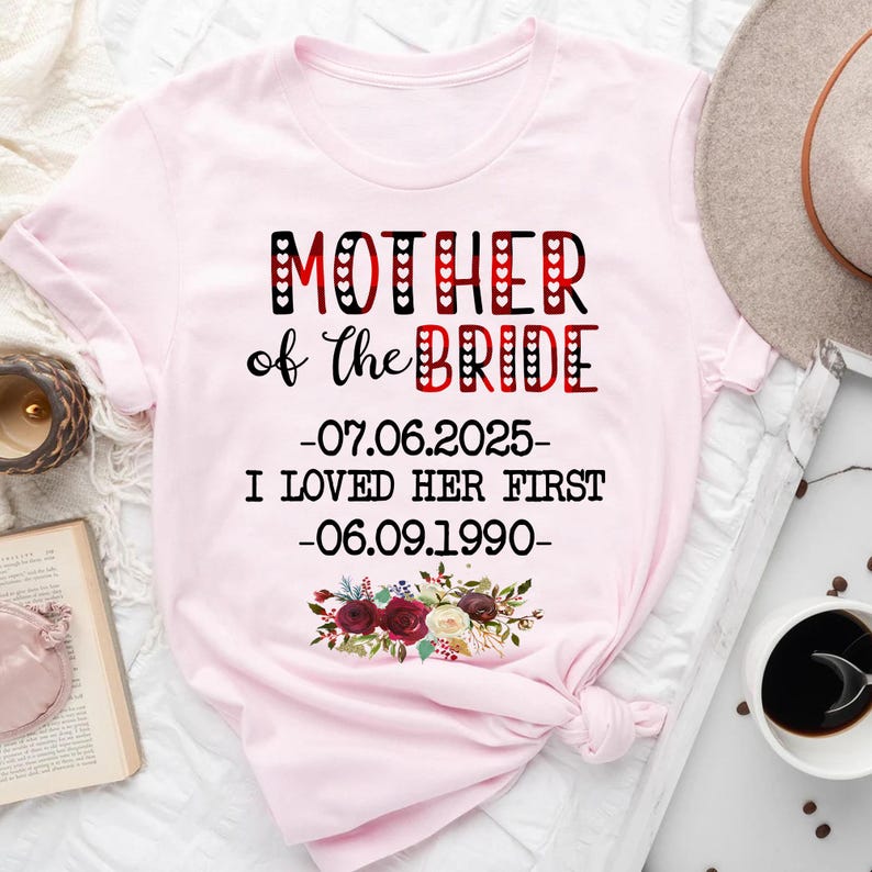 May include: Pink t-shirt with the text "Mother of the Bride - 07.06.2025 - I loved her first - 06.09.1990" in black and red with a floral design.