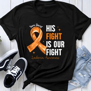 Könnte beinhalten: Ein schwarzes T-Shirt mit einer orangefarbenen Schleife und dem Text Team David HIS FIGHT IS OUR FIGHT Leukemia Awareness. Das Design enthält kleine orangefarbene Sternakzente. Das T-Shirt wird mit schwarz-weißen Turnschuhen und Jeans gezeigt.