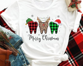 Op de afbeelding: Wit T-shirt met een kerstthema ontwerp met drie tanden die verkleed zijn als een rendier, elf en kerstman. De tekst "Merry Christmas" is onder de tanden gedrukt.
