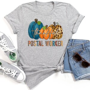 Puede incluir: Una camiseta gris claro con un diseño de temática otoñal. El diseño incluye tres calabazas en verde azulado, naranja y estampado de leopardo, con la palabra "thankful" en cursiva. Debajo de las calabazas, se muestra el texto "POSTAL WORKER".