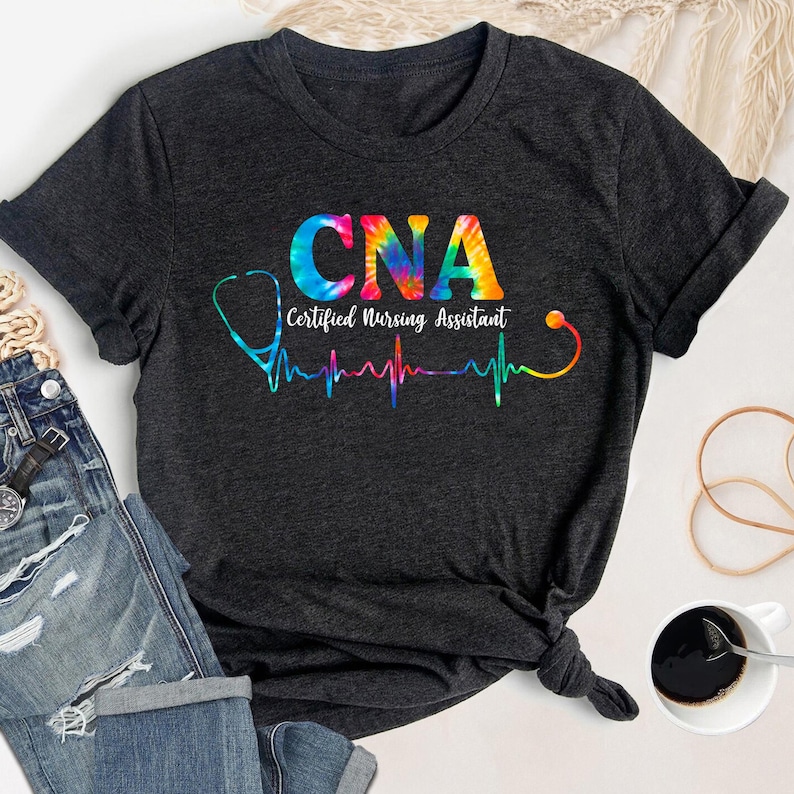 Puede incluir: Una camiseta gris oscuro con un dise&ntilde;o tie-dye que presenta el texto "CNA Certified Nursing Assistant" y un estetoscopio con un gr&aacute;fico de latido del coraz&oacute;n.