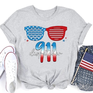 Puede incluir: Camiseta gris con un diseño de bandera estadounidense roja, blanca y azul. El diseño incluye gafas de sol con estrellas y rayas, el número 911 y la palabra "dispatcher".