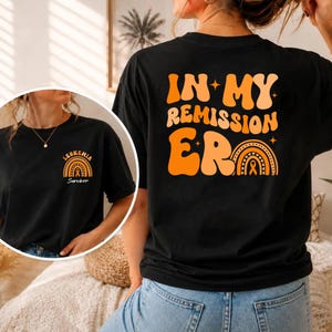 Könnte beinhalten: Ein schwarzes T-Shirt mit einer großen orangefarbenen und pfirsichfarbenen Grafik auf der Rückseite mit dem Text "IN MY REMISSION ERA" und einem Regenbogen mit Schleife. Die Brust zeigt eine kleinere passende Grafik mit "LEUKEMIA SURVIVOR".