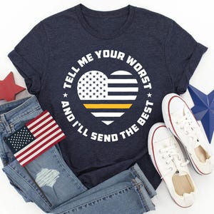 May include: A navy blue t-shirt with a white circle design featuring a heart-shaped American flag with a yellow stripe. The text "Tell me your worst and I'll send the best" is written around the heart.