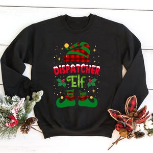 Könnte beinhalten: Schwarzes Sweatshirt mit einem festlichen "Dispatcher Elf" Design. Das Design zeigt einen rot-grün karierten Elfenhut, die Worte "Dispatcher Elf" in Rot und Grün und Elfenschuhe. Das Design ist mit Stechpalmen und Sternen verziert.