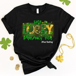 May include: Black t-shirt with a green, gold, and black graphic that says "One Lucky Pharmacy Tech" with a shamrock, horseshoe, and leprechaun hat. The text "#Your Hashtag" is below the graphic.