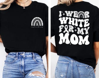 Puede incluir: Camiseta negra con un diseño de arcoíris y cinta blanca, con el texto "Concienciación sobre el cáncer de pulmón" en la parte delantera. La parte trasera de la camiseta presenta el texto "I wear white for my mom" en una fuente retro. La camiseta se combina con vaqueros azules.