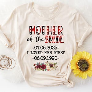 May include: A beige t-shirt with the text "Mother of the Bride" in red and black lettering. The text also includes the dates "07.06.2025" and "06.09.1990" and the phrase "I loved her first". The shirt has a floral design with red, white, and green flowers.
