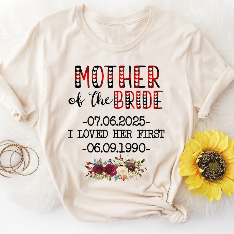 May include: A beige t-shirt with the text "Mother of the Bride" in red and black lettering. The text also includes the dates "07.06.2025" and "06.09.1990" and the phrase "I loved her first". The shirt has a floral design with red, white, and green flowers.