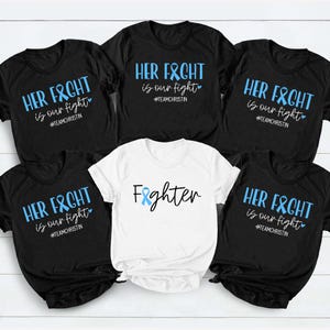 May include: A collection of black and white t-shirts with blue text and ribbon graphics. The shirts display the phrases "HER FIGHT is our fight" and "Fighter", along with the hashtag #TEAMCHRISTIN.