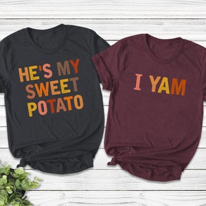 以下が含まれることがあります： 感謝祭をテーマにしたカップルのデザインが施されたTシャツ2枚。黒いTシャツには、オレンジ、茶色、黄色の文字で「He's my sweet potato」と書かれている。マルーンのTシャツには、オレンジ、茶色、黄色の文字で「I yam」と書かれている。