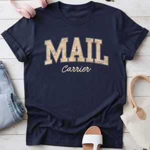 Könnte beinhalten: Ein dunkelblaues Kurzarm-T-Shirt mit dem Wort MAIL in großen Blockbuchstaben, die mit einem Muster aus alten Briefmarken und Umschlägen gefüllt sind, und dem Wort Carrier in einer helleren Schreibschrift darunter.