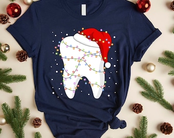 May include: A navy blue t-shirt featuring a graphic of a white tooth wearing a red Santa hat and wrapped in colorful Christmas string lights, surrounded by small white dots representing snow against the dark fabric background.
