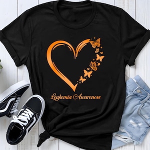 Könnte beinhalten: Ein schwarzes Kurzarm-T-Shirt mit einer orangefarbenen Grafik, die eine Herzumrisslinie zeigt, welche auf der rechten Seite aus mehreren Schmetterlingen besteht. Der Text "Leukemia Awareness" steht in orangefarbener Schreibschrift darunter.