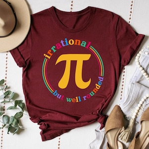 Peut inclure: Un t-shirt marron avec un graphique du symbole mathématique pi dans un cercle avec le texte "irrational but well rounded" autour. Le texte est en couleurs arc-en-ciel.