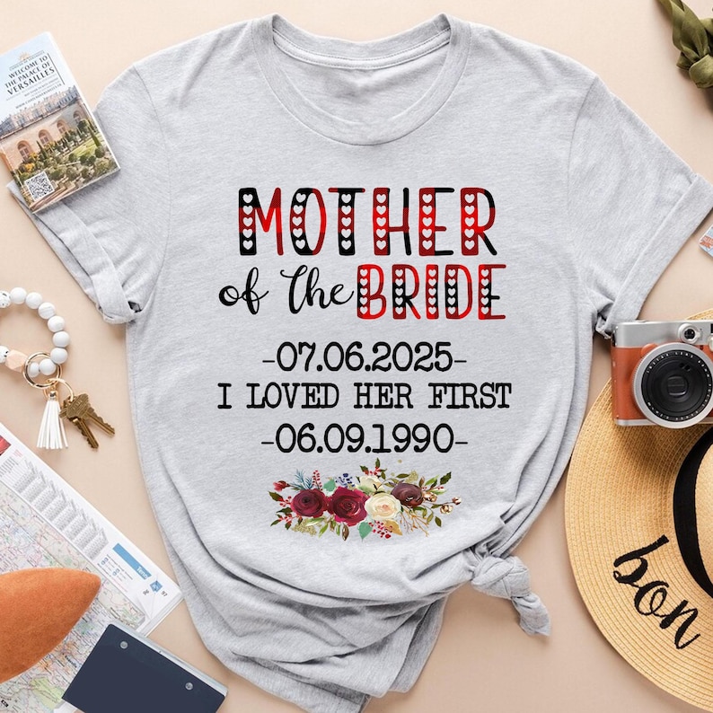 May include: Gray t-shirt with red and white plaid design. The text "Mother of the Bride" is printed in black with red hearts. The dates "07.06.2025" and "06.09.1990" are printed below the text, along with the phrase "I loved her first".