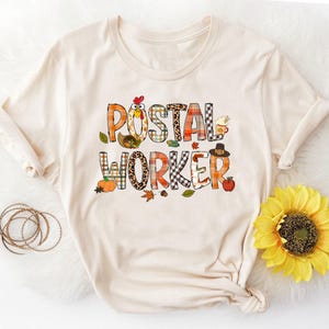 Könnte beinhalten: Ein beiges T-Shirt mit einem Thanksgiving-Motiv, auf dem "Postangestellter" steht. Das Design zeigt einen Truthahn, Kürbisse, Blätter und Karomuster.