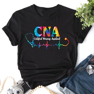 Puede incluir: Camiseta negra con un dise&ntilde;o tie-dye de las letras CNA, que significa Certified Nursing Assistant. Las letras est&aacute;n rodeadas de un estetoscopio y un gr&aacute;fico de latido del coraz&oacute;n.