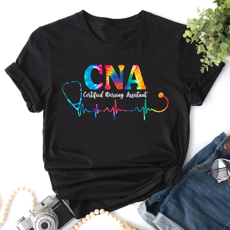 Puede incluir: Camiseta negra con un dise&ntilde;o tie-dye de las letras CNA, que significa Certified Nursing Assistant. Las letras est&aacute;n rodeadas de un estetoscopio y un gr&aacute;fico de latido del coraz&oacute;n.