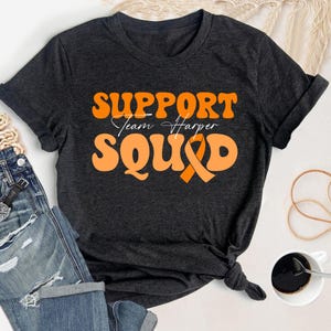 Può includere: T-shirt grigio scuro con la scritta arancione "SUPPORT Team Harper SQUAD". Un nastro arancione di sensibilizzazione è incorporato nel design. La maglietta è annodata in basso.