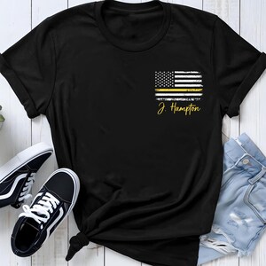 May include: A black short-sleeve t-shirt featuring a distressed American flag graphic on the left chest area. The flag includes a horizontal yellow stripe and the text J. Hampton printed in yellow cursive script directly below the flag design.