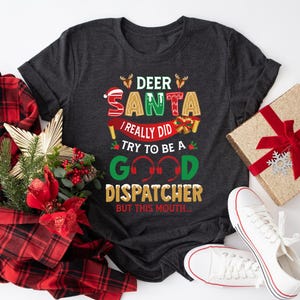Op de afbeelding: Een donkergrijs T-shirt met een kerstthema. Het ontwerp bevat een rood-groene kerstmanmuts, een peperkoekman en de tekst "Deer Santa I really did try to be a good dispatcher but this mouth..."