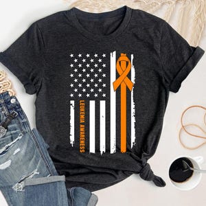 Può includere: T-shirt grigio scuro con un disegno della bandiera americana, un nastro arancione e la scritta "LEUKEMIA AWARENESS". La bandiera ha strisce e stelle bianche. Il nastro arancione è un simbolo di consapevolezza della leucemia.