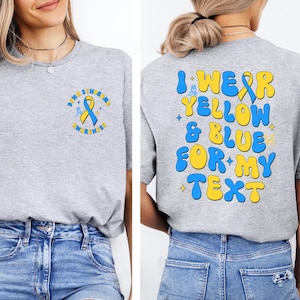 Op de afbeelding: Een grijs gemêleerd t-shirt met een klein Downsyndroom bewustzijnslintje op de linkerborst. De achterkant toont de tekst I WEAR YELLOW & BLUE FOR MY TEXT in grote, dikke, golvende blauwe en gele letters met kleine sterretjes als accent.
