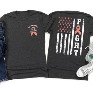 May include: Dark grey t-shirts featuring a support ribbon design. One shirt displays "SUPPORT SQUAD" and "TEAM NAME". The other has a flag design with the word "FIGHT" and ribbons.