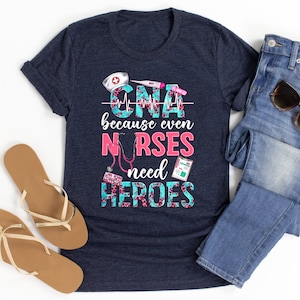 Puede incluir: Una camiseta azul marino con un diseño gráfico rosa y azul que dice "CNA because even NURSES need HEROES". El gráfico incluye un estetoscopio, un gorro de enfermera, un termómetro y una botella de pastillas.