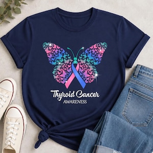 May include: A navy blue t-shirt featuring a large butterfly graphic composed of smaller pink, purple, and teal butterflies. A split pink and blue awareness ribbon forms the butterfly body. The text Thyroid Cancer AWARENESS is printed below.