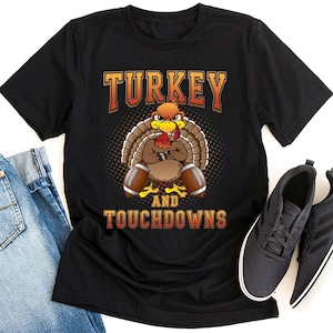 May include: Black t-shirt featuring a cartoon turkey with a football and the text "TURKEY AND TOUCHDOWNS" in orange and brown. The turkey is brown with a red wattle and yellow feet. The shirt is paired with dark gray sneakers and blue jeans.