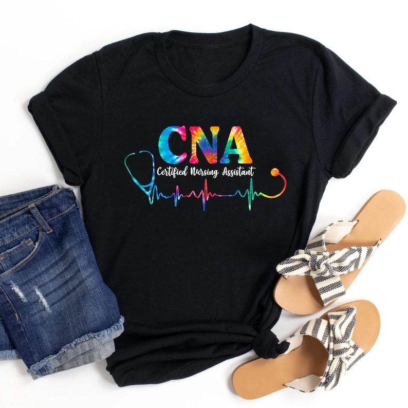 Puede incluir: Camiseta negra con un dise&ntilde;o tie-dye que presenta el texto "CNA Certified Nursing Assistant" y un estetoscopio con un gr&aacute;fico de latido del coraz&oacute;n.