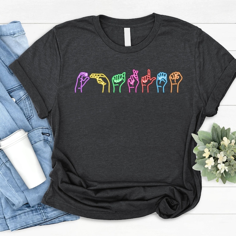 Op de afbeelding: Een donkergrijs T-shirt met een regenboogontwerp van handen die het woord "LIEfde" in Nederlandse Gebarentaal tekenen.