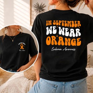 Può includere: Una t-shirt nera con una grafica sul retro che recita IN SEPTEMBER WE WEAR ORANGE in caratteri ondulati arancioni e bianchi, con sotto Leukemia Awareness. La zona del petto mostra un piccolo nastro arancione e il testo SUPPORT SQUAD.