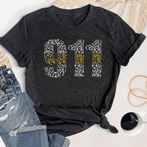 May include: A dark grey t-shirt with a white and yellow graphic design of the number 911. The number is made up of small circles with a yellow outline.