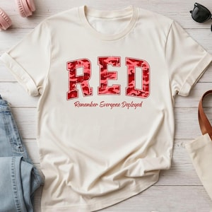 Könnte beinhalten: Ein cremefarbenes Kurzarm-T-Shirt mit dem Wort RED in großen Blockbuchstaben mit rotem Tarnmuster. Unter dem Wort ist der Text Remember Everyone Deployed in einer roten, kursiven Schriftart aufgedruckt.