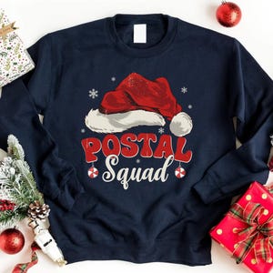 Könnte beinhalten: Marineblaues Sweatshirt mit einem Weihnachtsmützen-Motiv und dem Text "POSTAL Squad" in Rot und Weiß. Das Design umfasst Schneeflocken und Ornamente, ideal für Postangestellte in der Weihnachtszeit.