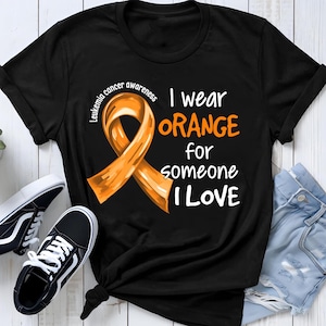 Può includere: T-shirt nero con un nastro arancione di sensibilizzazione e la scritta "I wear ORANGE for someone I LOVE." La maglietta include anche le parole "Leukemia cancer awareness." Sono visibili anche scarpe da ginnastica nere e bianche e jeans blu.