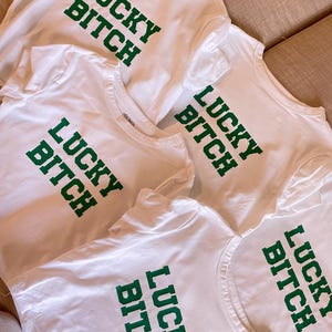 Può includere: Quattro magliette bianche con la scritta "Lucky Bitch" stampata in verde sul davanti.